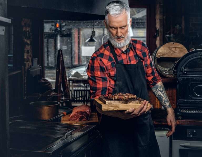 Tattooed elderly butcher holding board with bbq in meat shop